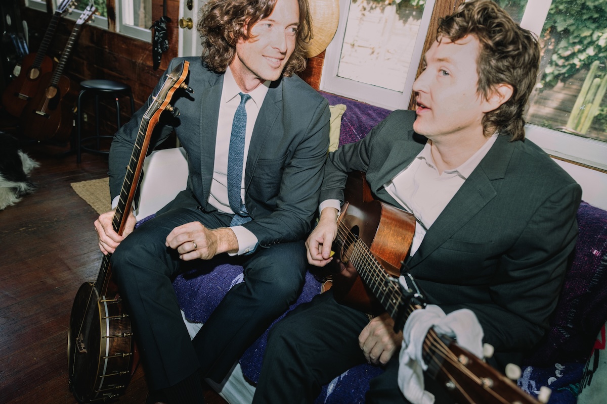 Folk duo The Milk Carton Kids sitting indoors in suits, holding a acoustic guitar and banjo while seated on a couch in a cozy studio setting.